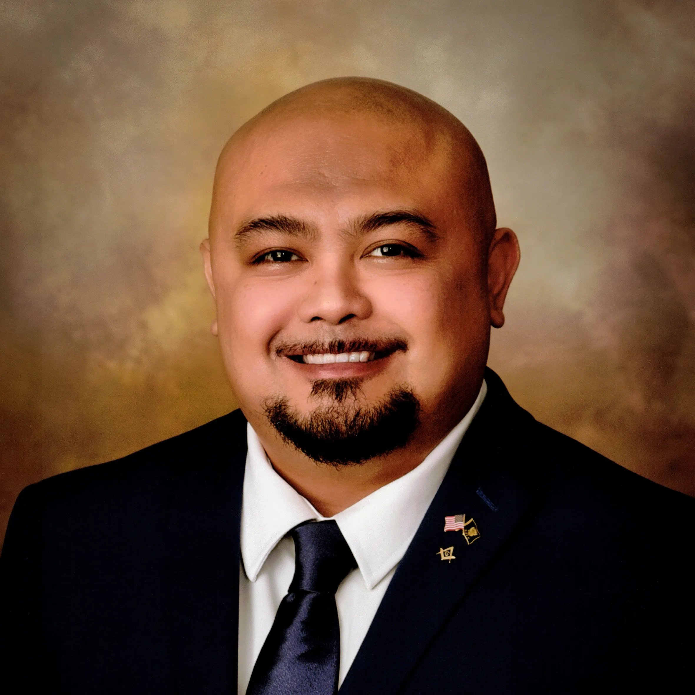 Jonathan Cruz in a dark suit and tie against a warm studio backdrop, wearing small lapel pins.